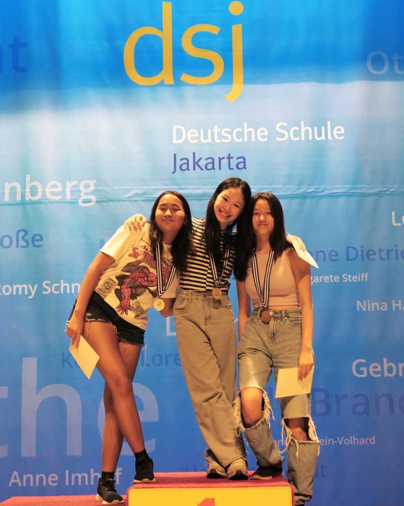 Three girls stand on a podium in front of a sign that says dsj