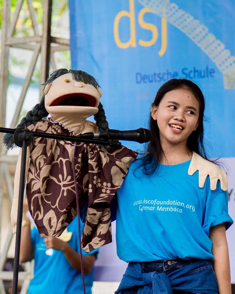 A girl wearing a blue shirt that says dsj stands next to a puppet