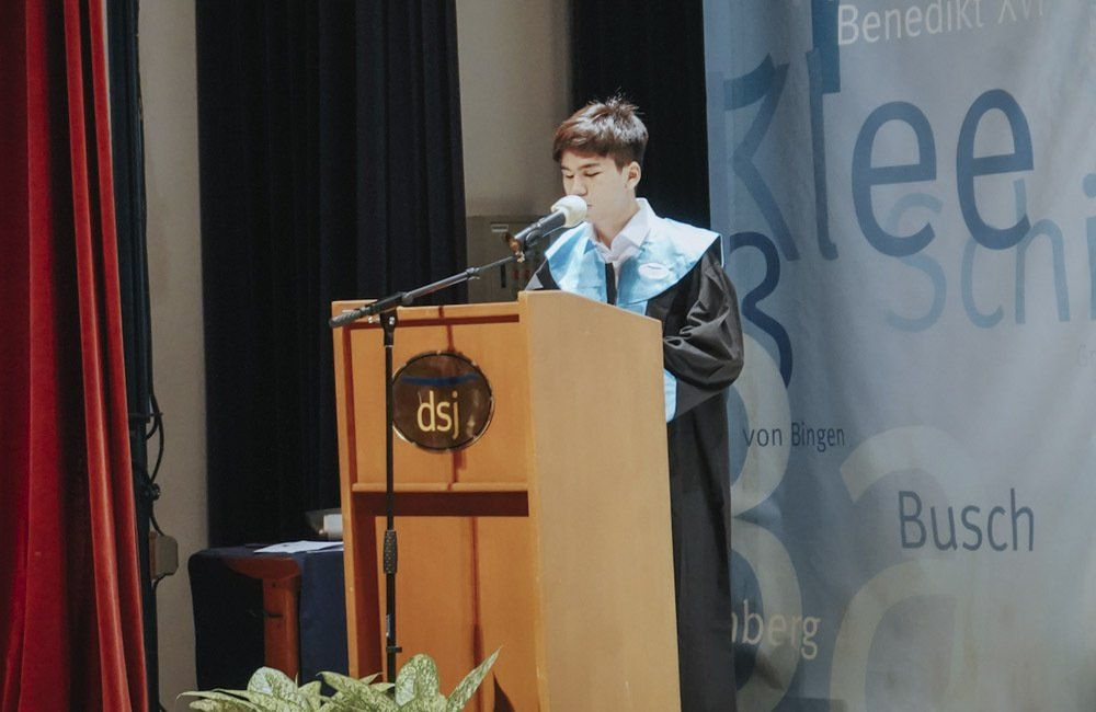 A man in a graduation gown is standing at a podium speaking into a microphone.
