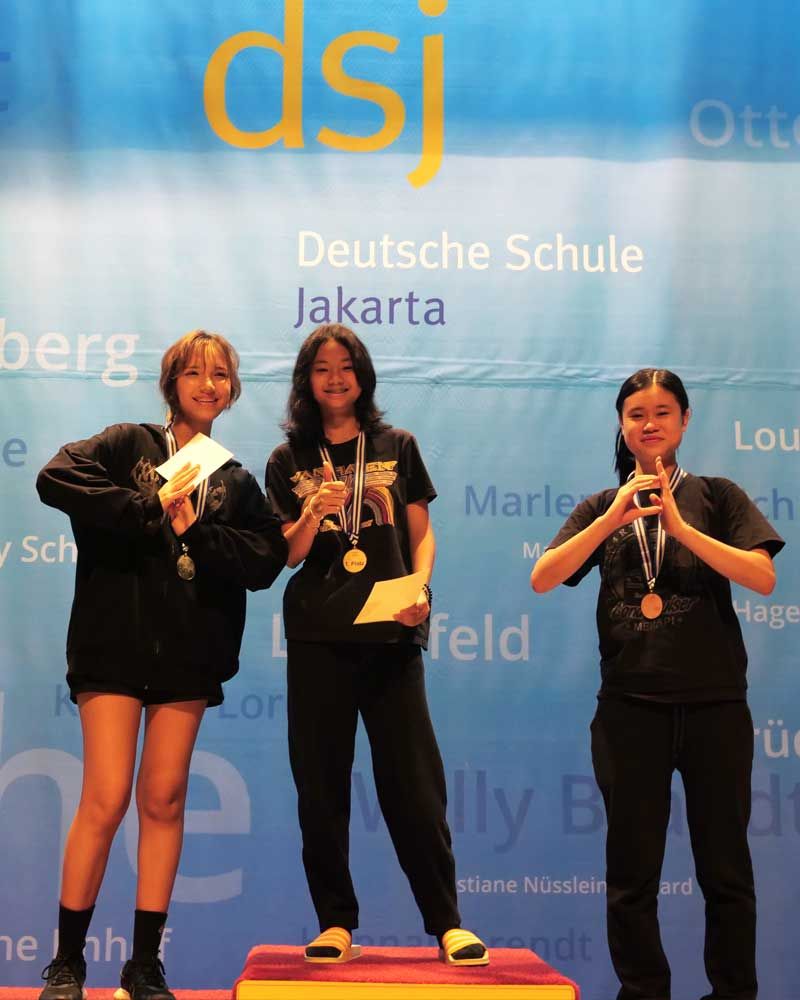 Three girls stand on a podium in front of a wall that says dsj