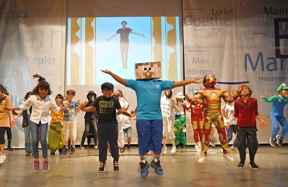 A group of children are dancing on a stage in front of a large screen.