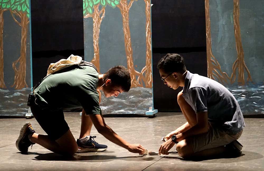 Two men are kneeling on a stage in front of a painting of trees.