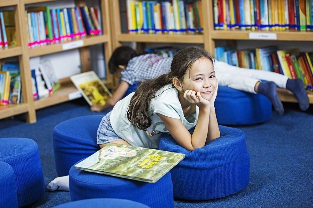 A girl is laying on a blue bean bag chair reading a book in a library.