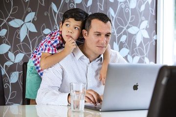 A man is holding a child on his shoulders while using a laptop computer.