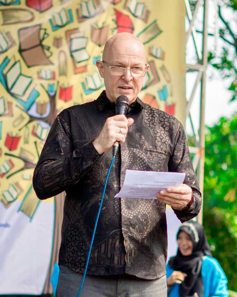A bald man is speaking into a microphone while holding a piece of paper