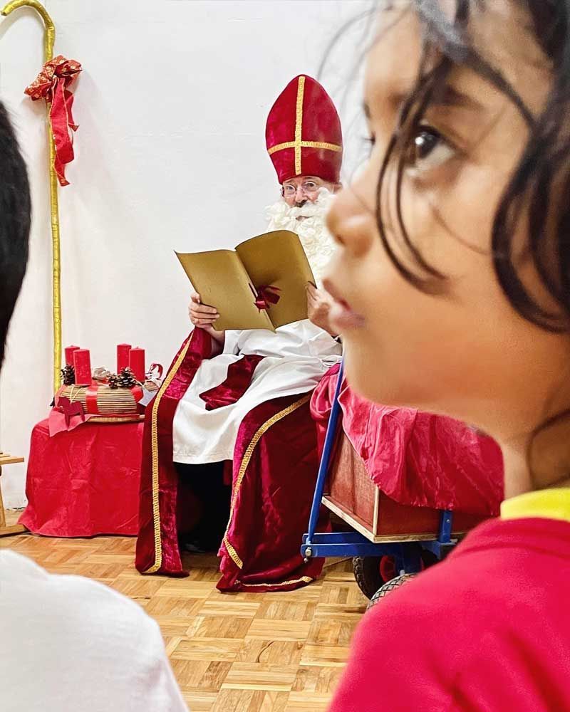 A little girl is looking at a man in a santa costume reading a book