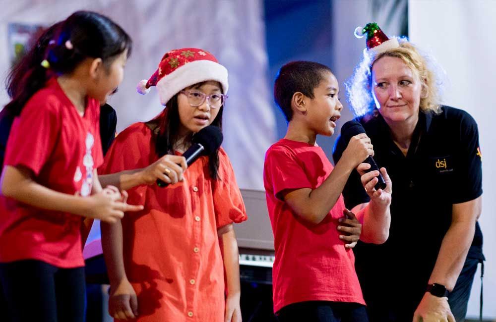 A group of children are singing into microphones while a woman stands behind them.