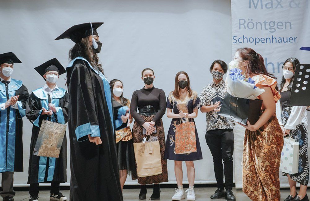 A woman in a graduation cap and gown is standing in front of a group of people wearing masks.
