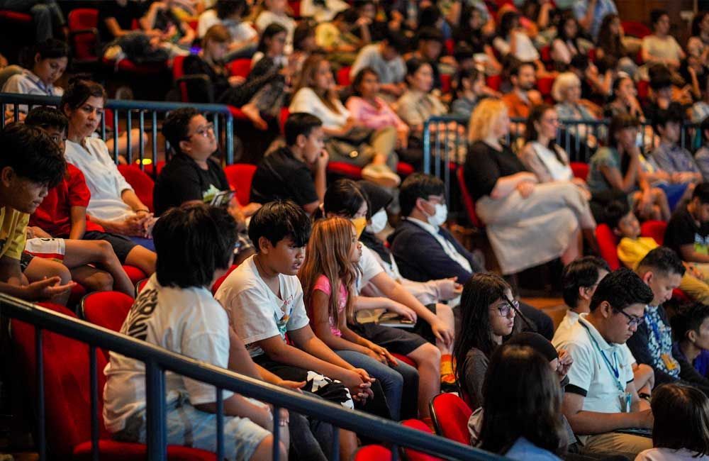 A large group of people are sitting in red chairs in an auditorium.