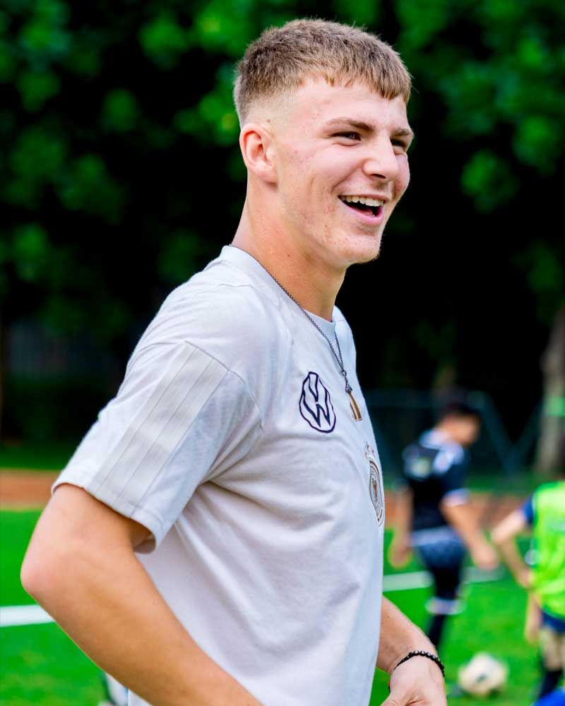 A young man in a white shirt is smiling on a soccer field.