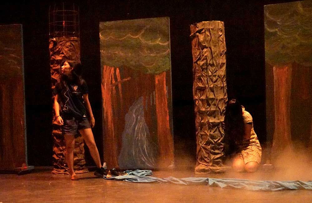 Two women are standing on a stage in front of a painting of trees.