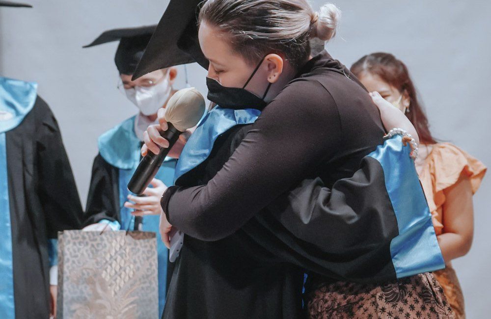 A woman in a graduation cap and gown is hugging another woman while holding a microphone.
