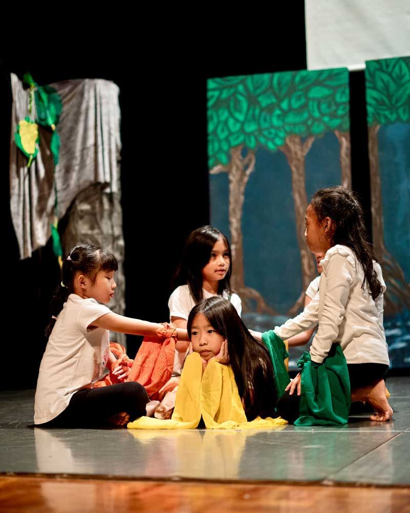 A group of young girls are sitting on the floor on a stage.