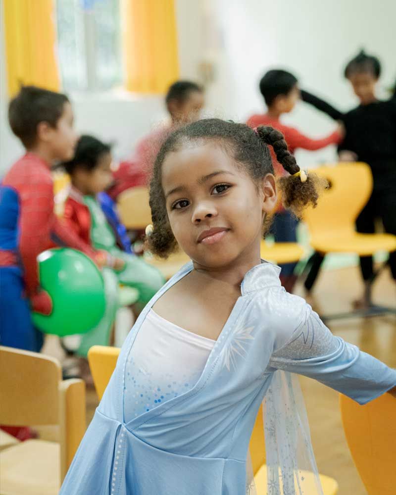A little girl in a blue dress is standing in front of a group of children.