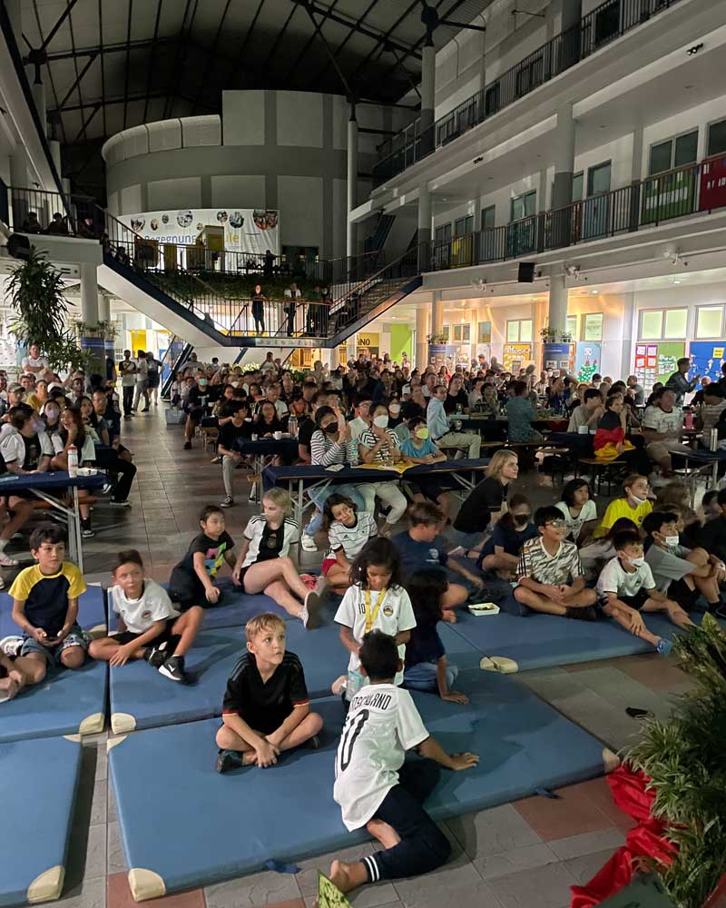 A large group of children are sitting on a blue mat in a gym.