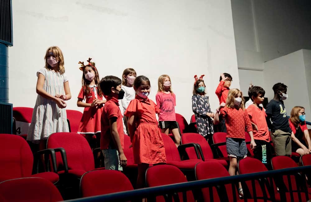 A group of children wearing face masks are standing in a row of red chairs.