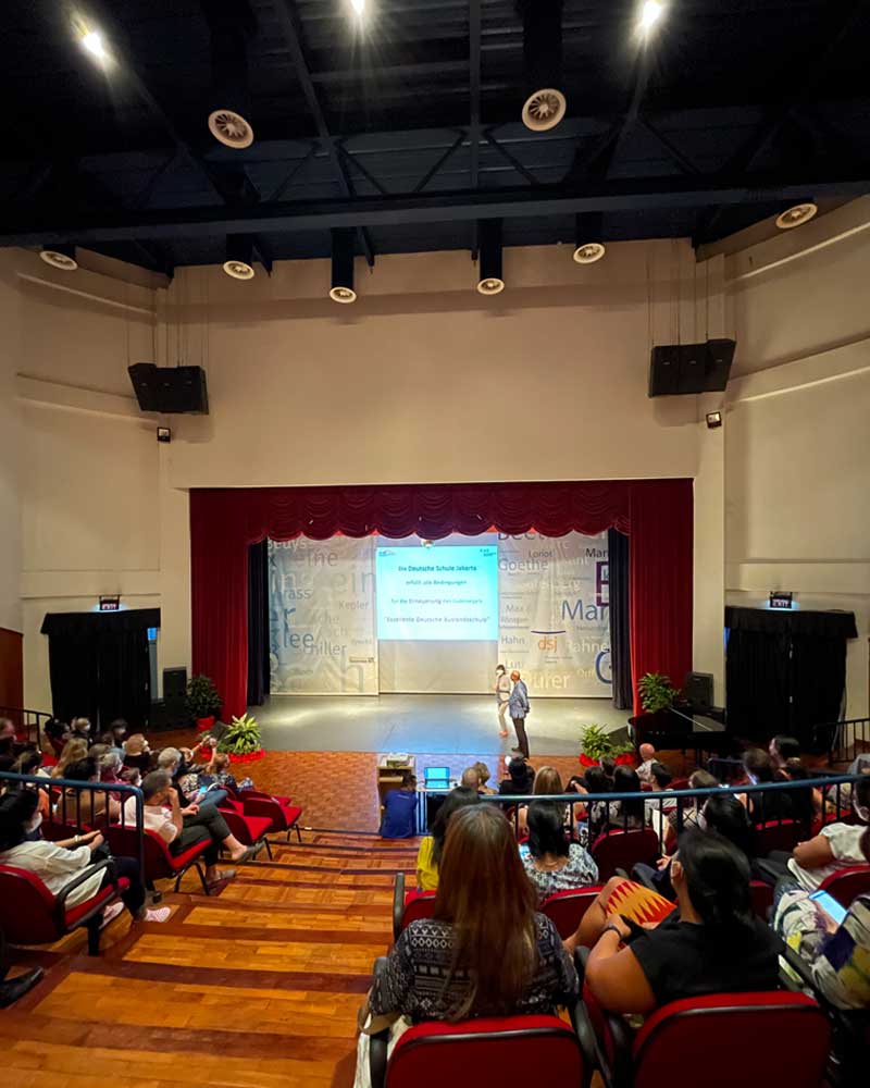 A group of people are sitting in an auditorium watching a presentation on a stage.