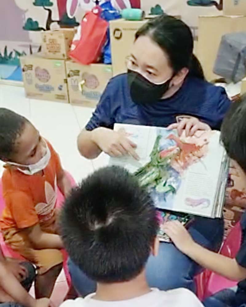 A woman wearing a mask is reading a book to a group of children.