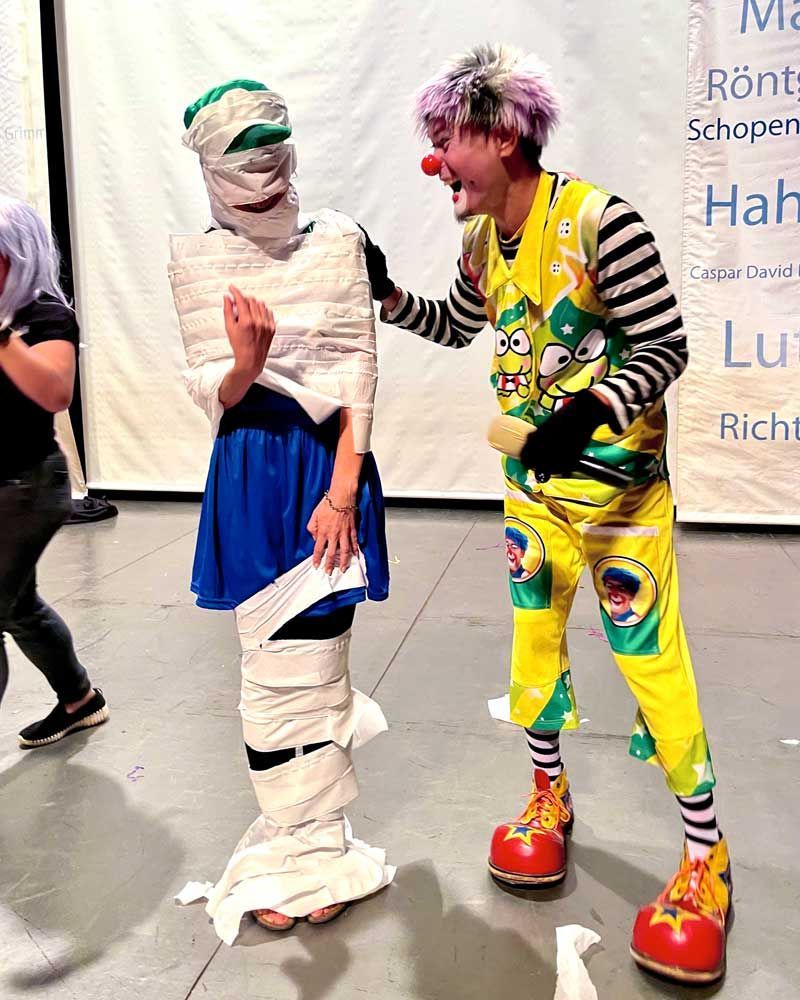 A clown is standing next to a mummy wrapped in toilet paper.