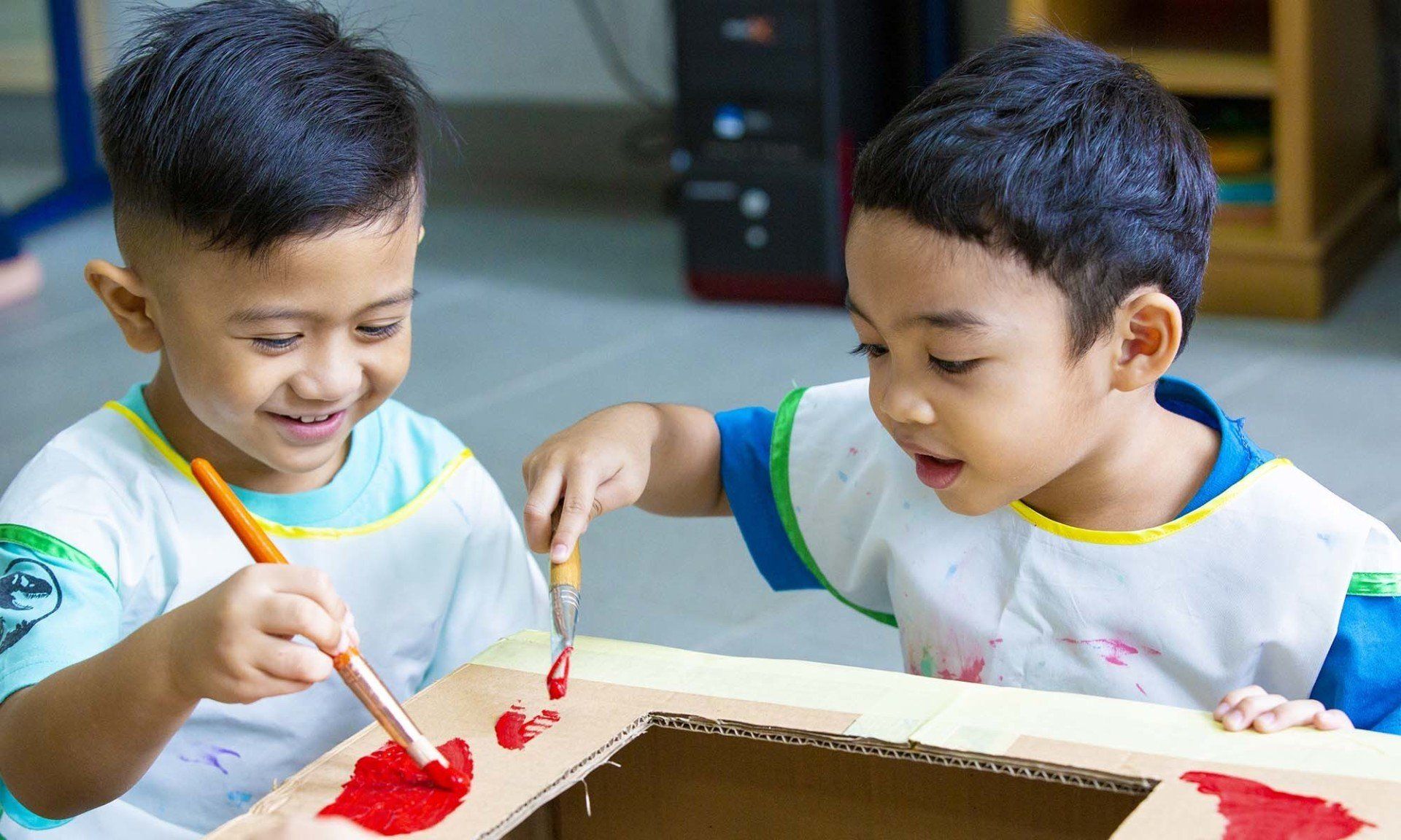 Two young boys are painting a cardboard box with red paint.