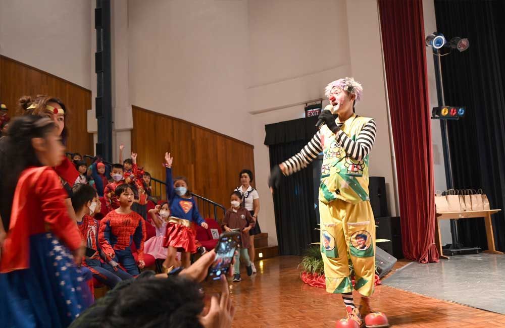 A clown is standing on a stage with a microphone in front of a crowd of children.