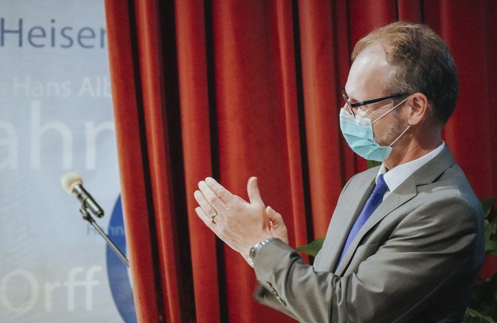 A man in a suit and tie wearing a face mask is applauding while standing in front of a microphone.