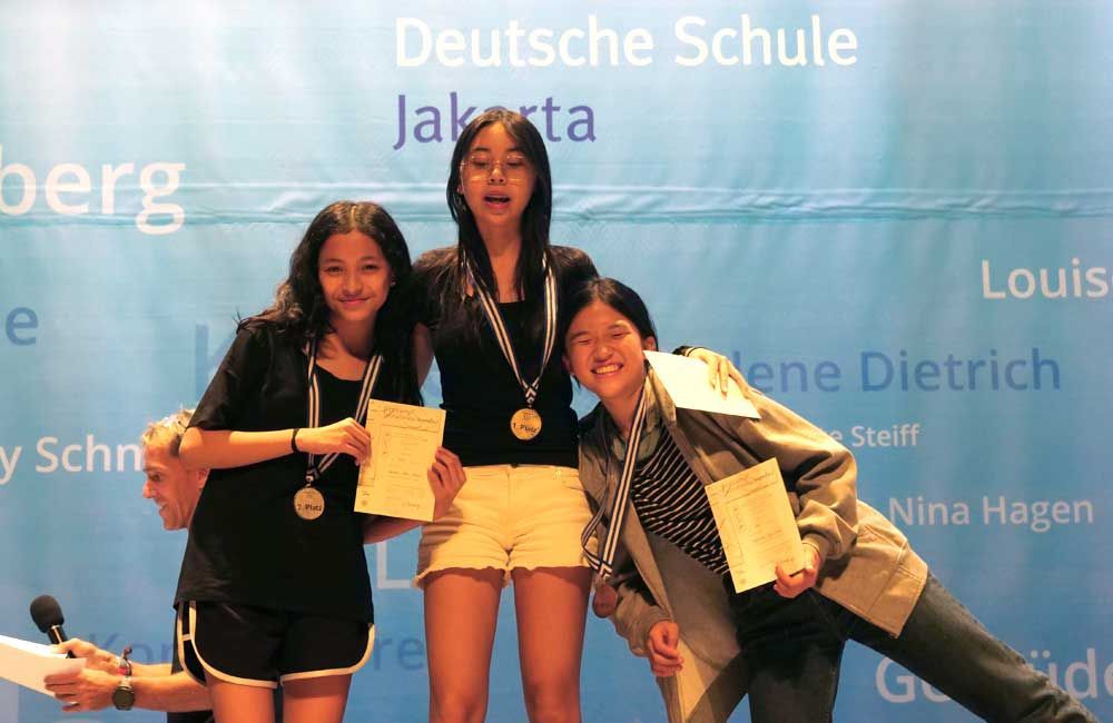 Three girls are posing for a picture in front of a wall that says deutsche schule
