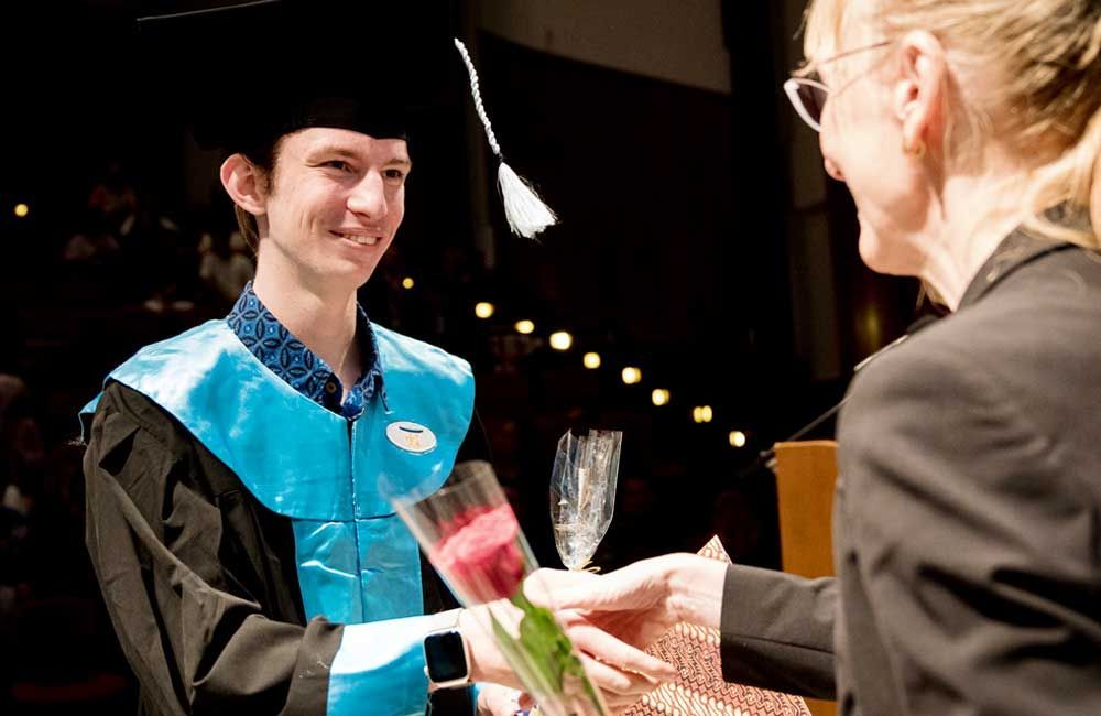 A man in a graduation cap and gown is receiving flowers from a woman