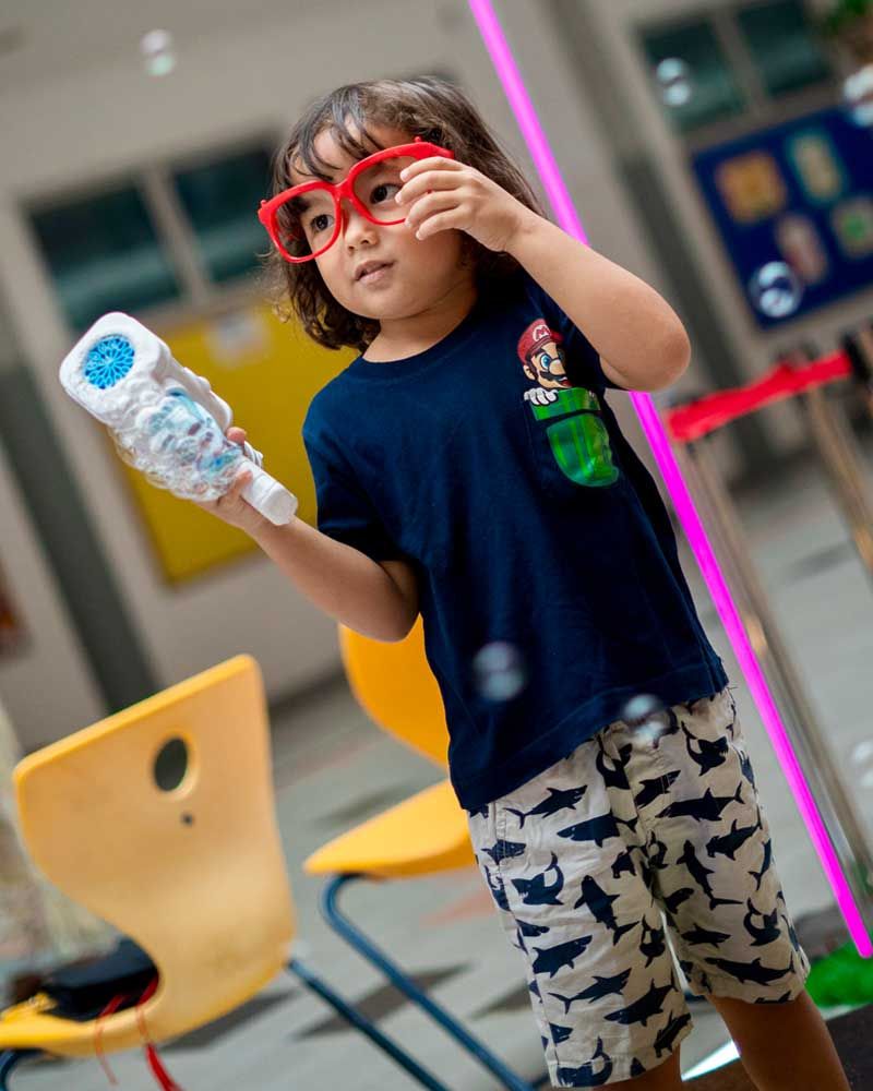 A little girl wearing glasses is blowing bubbles with a bottle.