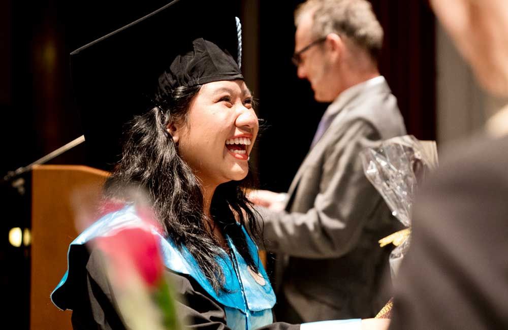 A woman in a graduation cap and gown is smiling while a man in a suit stands behind her.
