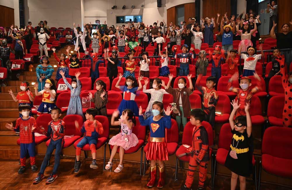 A large group of children dressed in superhero costumes are sitting in an auditorium.