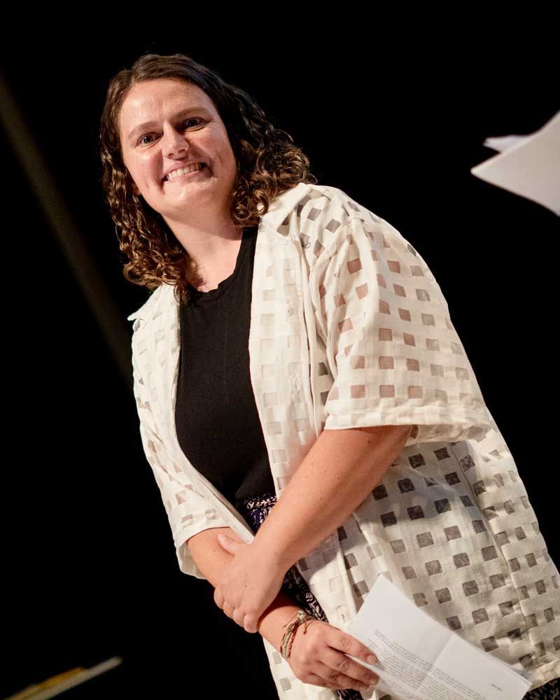 A woman wearing a black shirt and a white jacket is smiling and holding a piece of paper