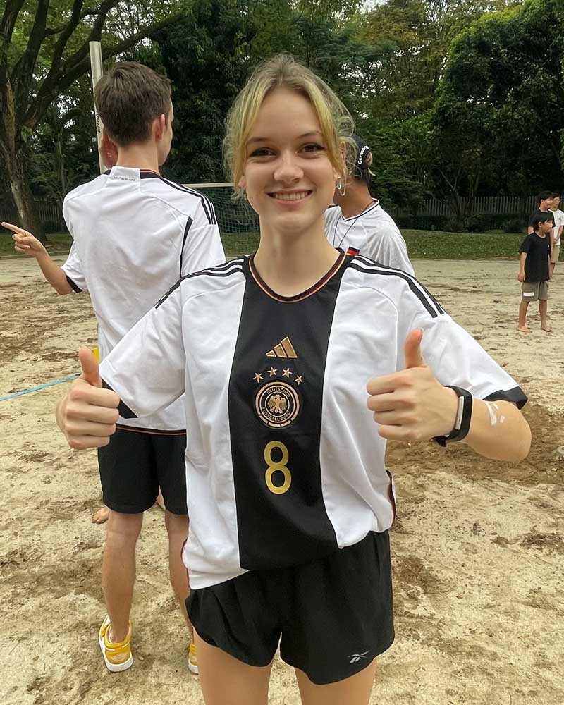 A young woman in a Germany soccer jersey smiles and gives a thumbs-up while standing on a sandy sports field.