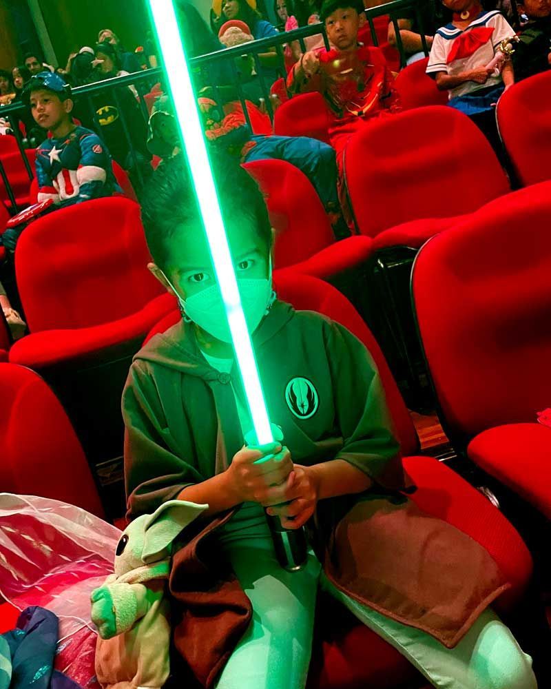 A young boy is holding a green lightsaber in a theater