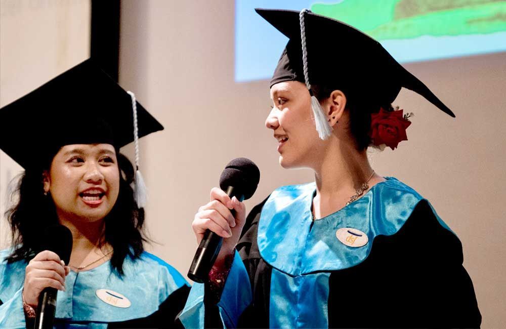 Two women in graduation caps and gowns are speaking into microphones