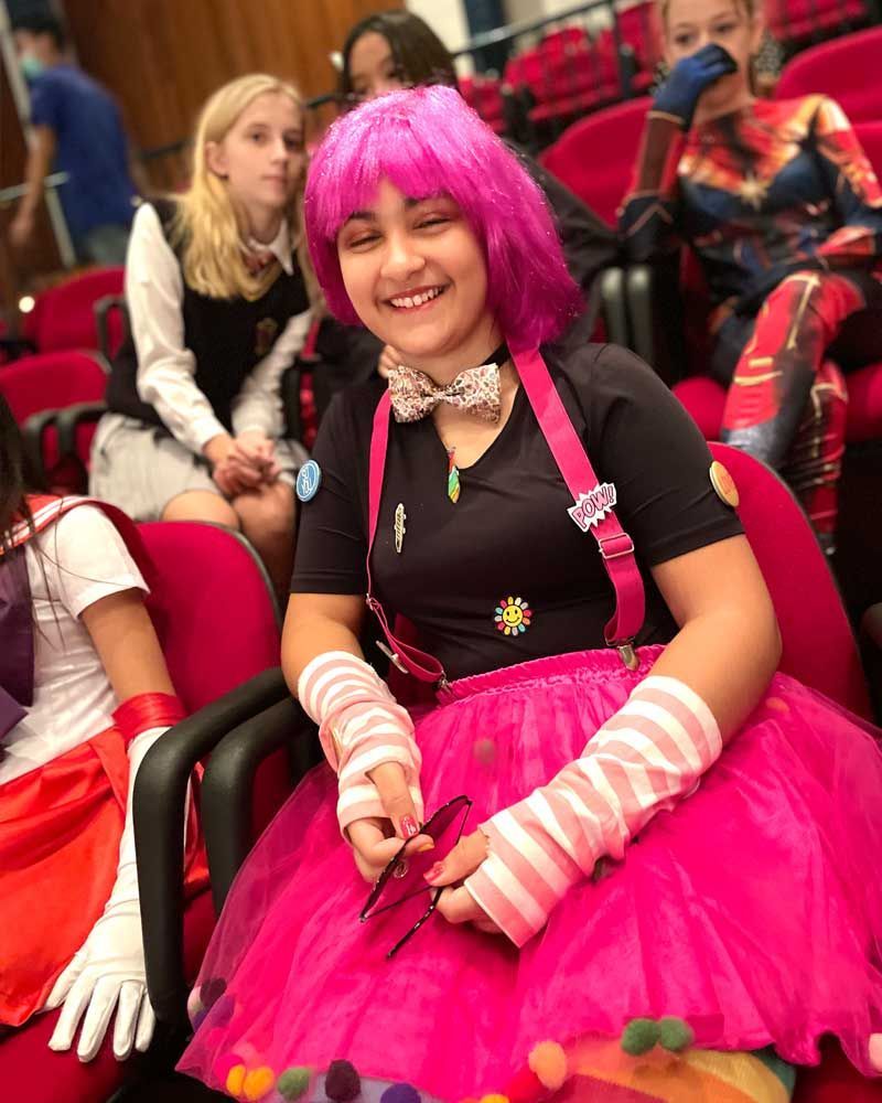A girl with pink hair is sitting in a chair holding a pair of scissors