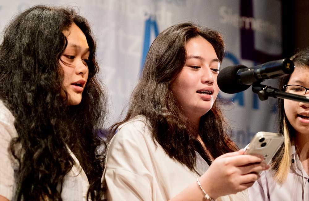 Three young women are singing into microphones while one of them is holding a cell phone.