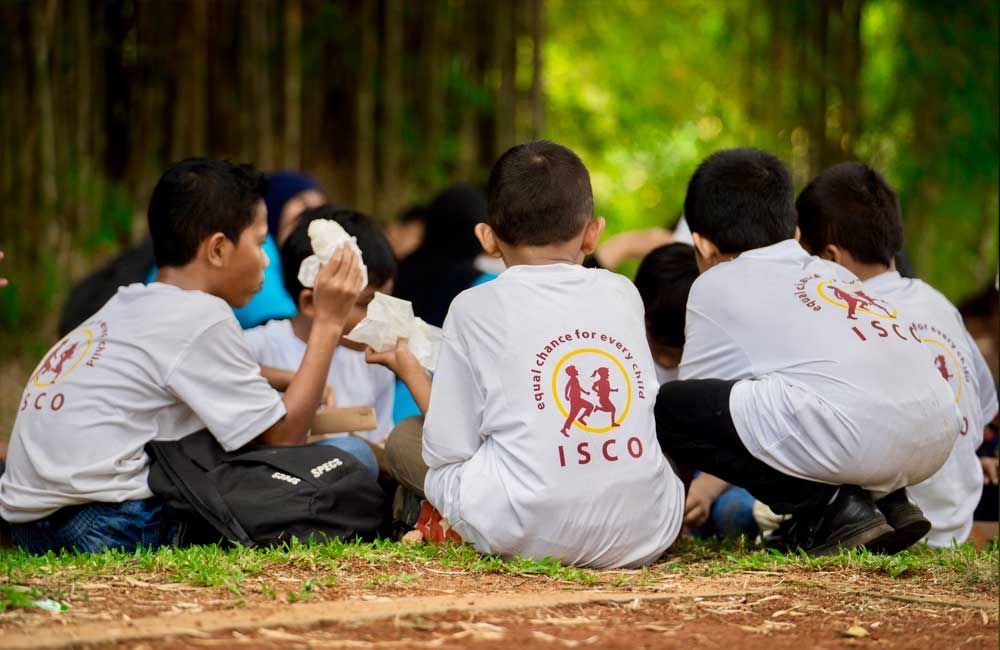A group of children wearing white shirts with the word isco on the back are sitting on the grass.