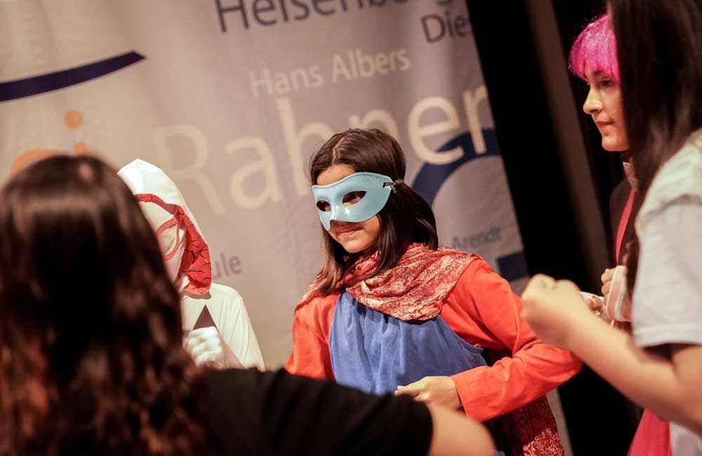 A woman wearing a mask stands in front of a sign that says hans albers