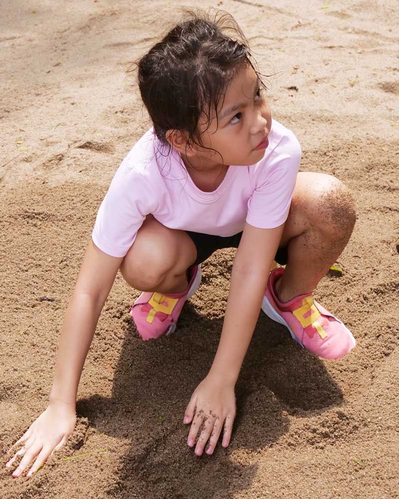 A little girl in a pink shirt and pink shoes is squatting in the sand