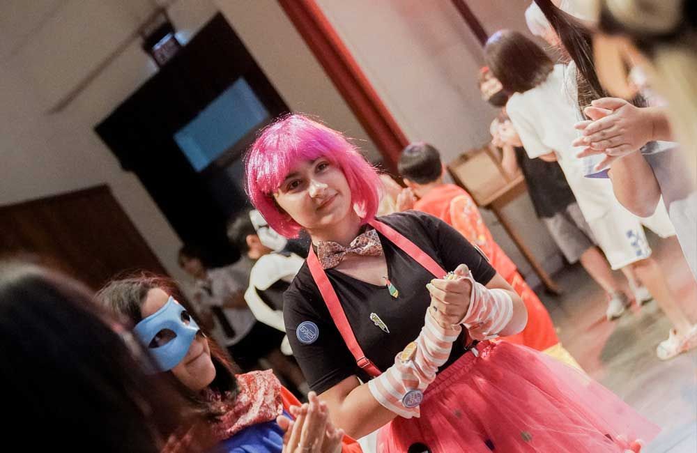 A woman with pink hair and a blue mask is standing in a room with other people.
