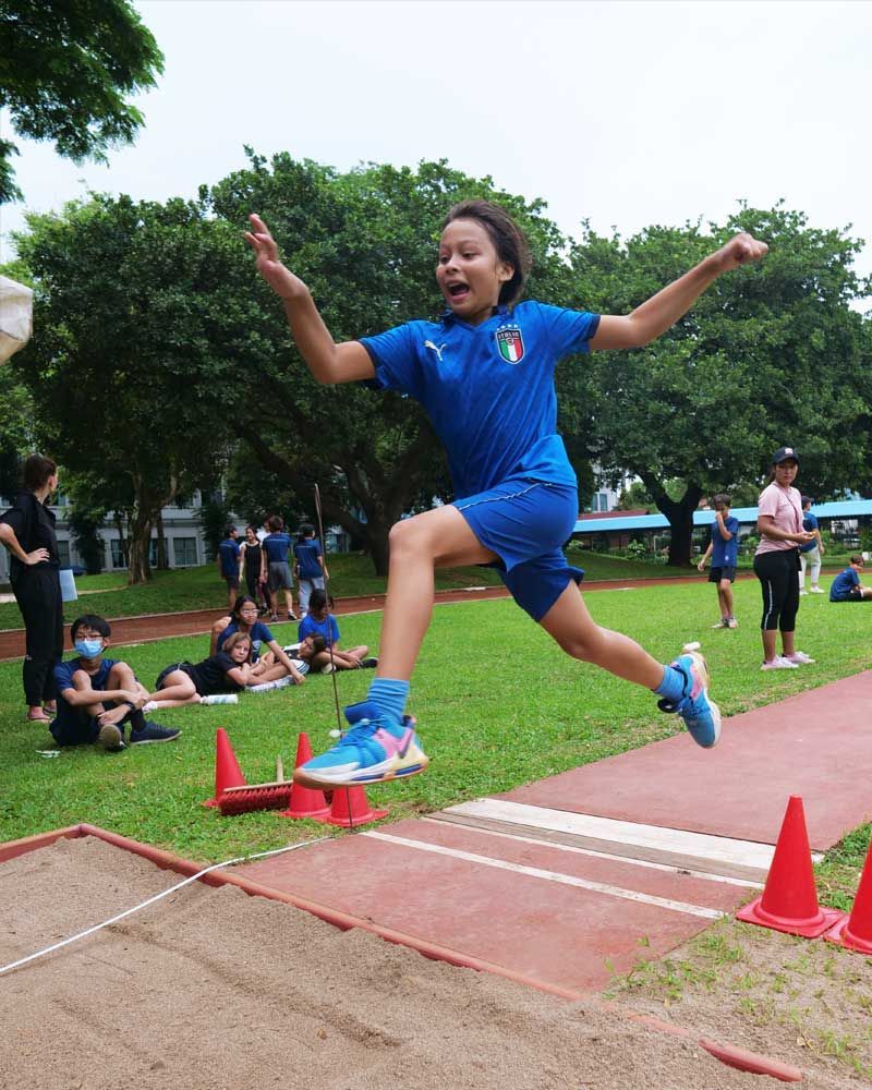 A young girl in a blue shirt is jumping in the air on a track.
