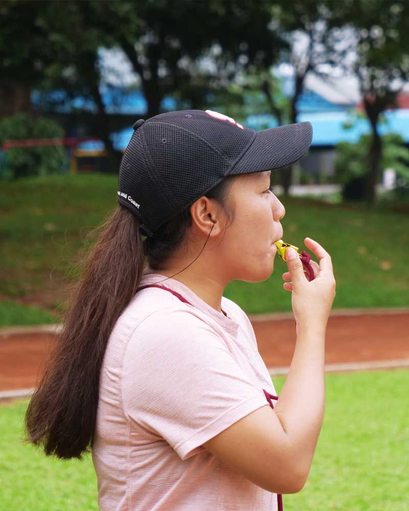 A woman wearing a baseball cap and a pink shirt is blowing a whistle.