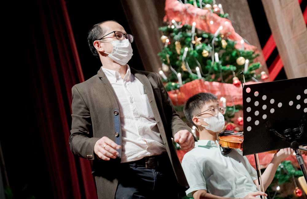 A man wearing a mask is standing next to a boy playing a violin in front of a christmas tree.
