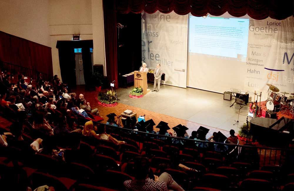 A large group of people are sitting in an auditorium watching a presentation on a stage.
