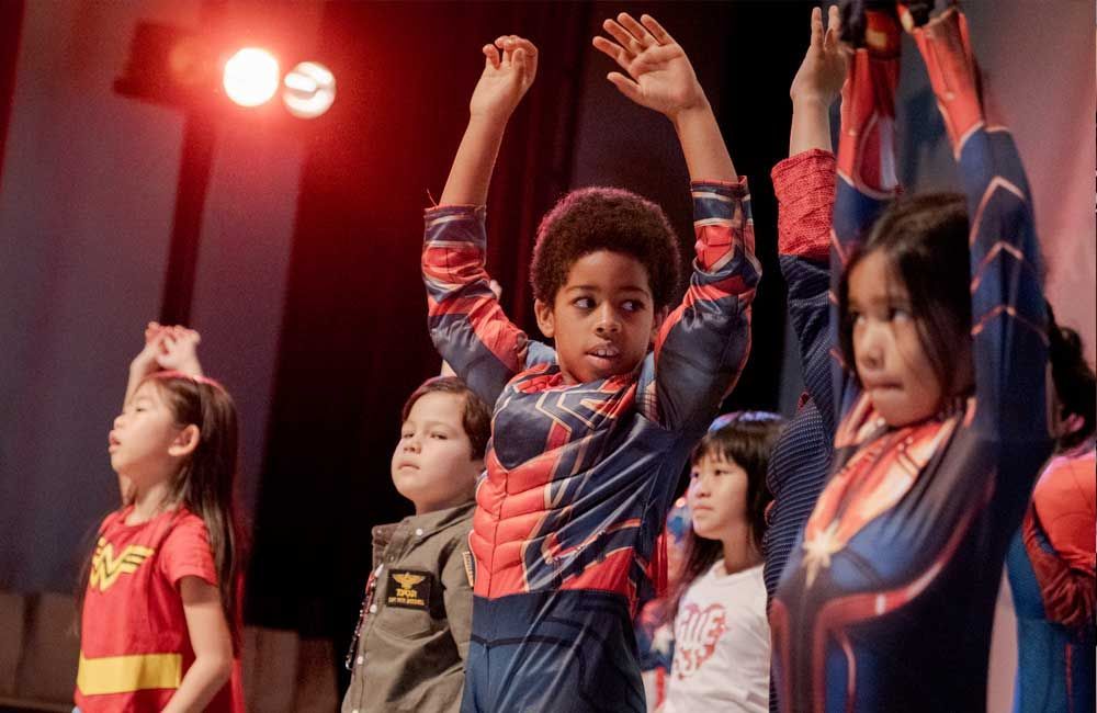 A group of children in superhero costumes are dancing on a stage.