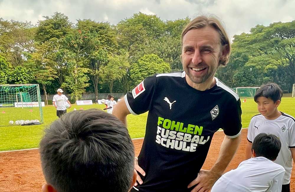 A man in a soccer uniform is talking to a group of children on a field.
