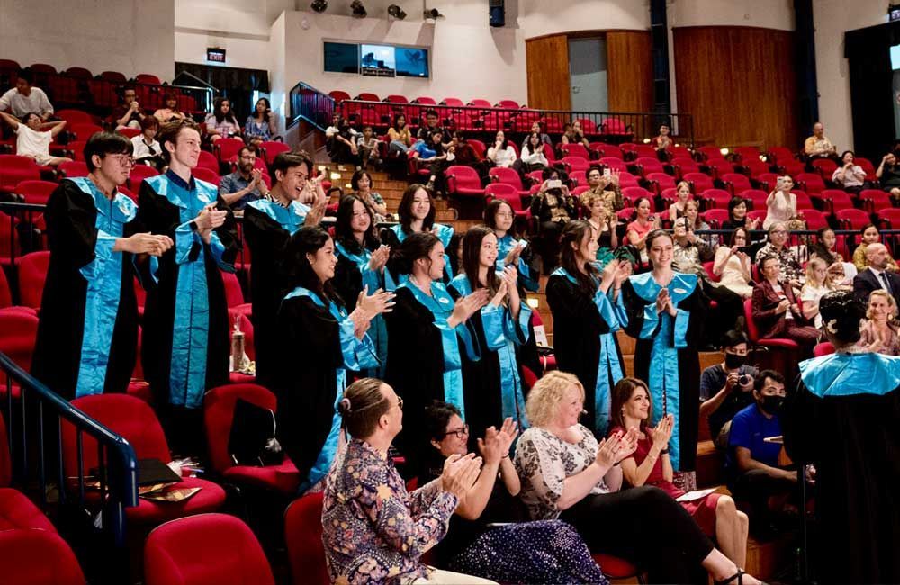 A group of people in graduation gowns are clapping in an auditorium.