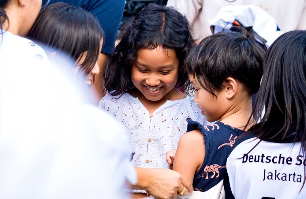 A group of children are standing around a girl wearing a shirt that says deutsche jakarta.