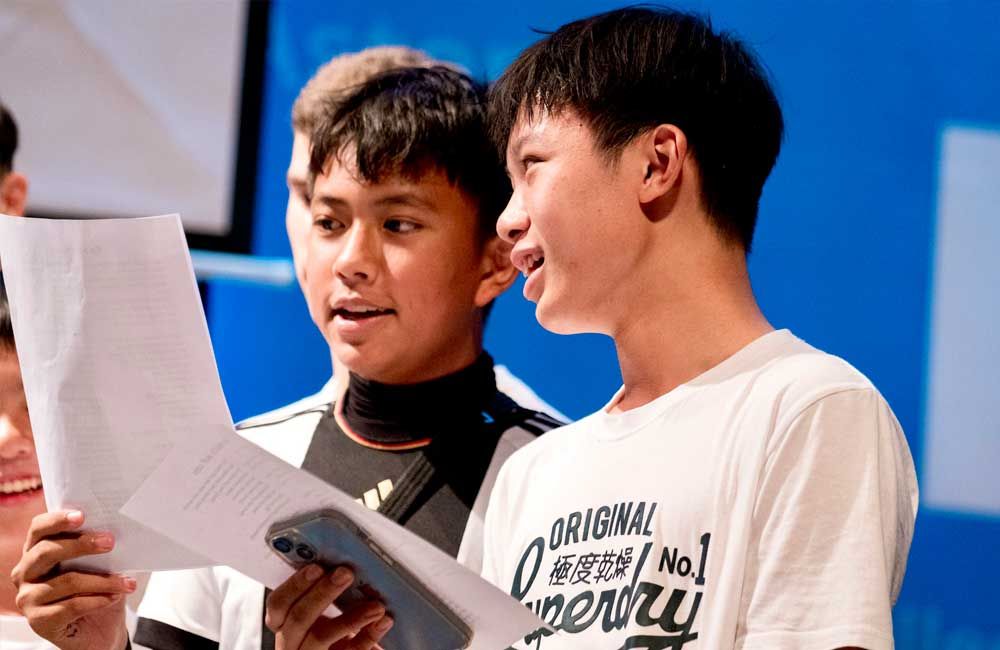 Two young boys are standing next to each other reading a piece of paper.